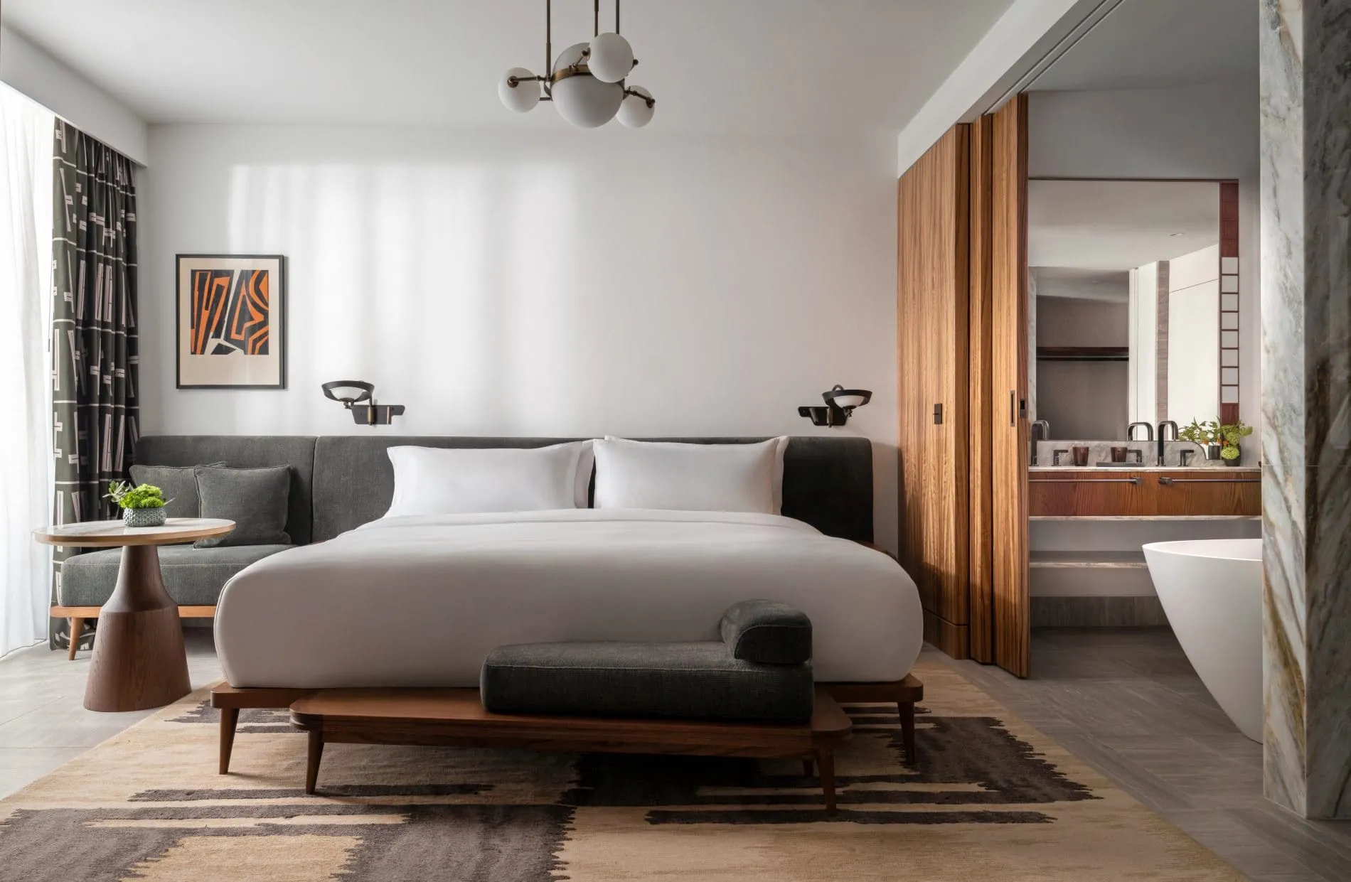 Die Deluxe Suite zählt zu den 278 Zimmern und Suiten, die laut Betreiber zu den größten Athens gehören. Mid-Century-Möbel, warme Holztöne und ein offen gestaltetes Marmorbad spiegeln die Designhandschrift des Studios Avroko wider.