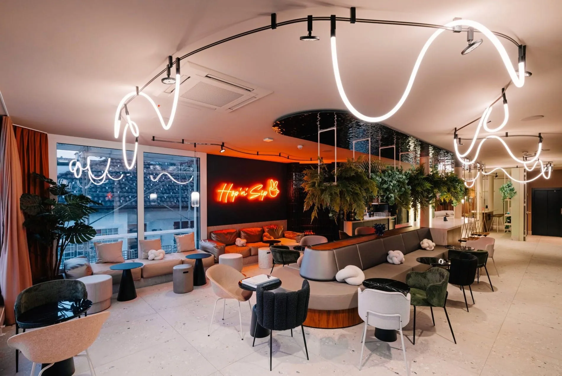Im Lounge-Bereich setzt das Studio Monoplan auf geschwungene Lichtlinien von Artemide, die mit dem orangefarbenen „Hop’n’Sip“-Schriftzug und üppigem Grün eine entspannte Atmosphäre zwischen Café und Wohnzimmer schaffen.