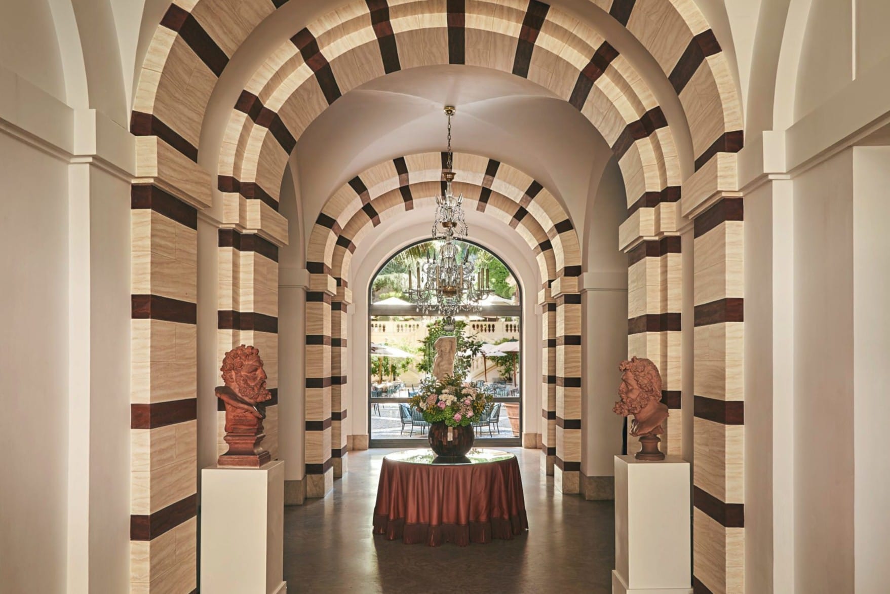 Klassische römische Rundbögen mit kontrastreichen Streifen rahmen den Blick in den „Secret Garden“ des Hotels de Russie. Antike Terrakotta-Büsten auf weißen Säulen und ein Kristallleuchter unterstreichen das Flair des Entrees.