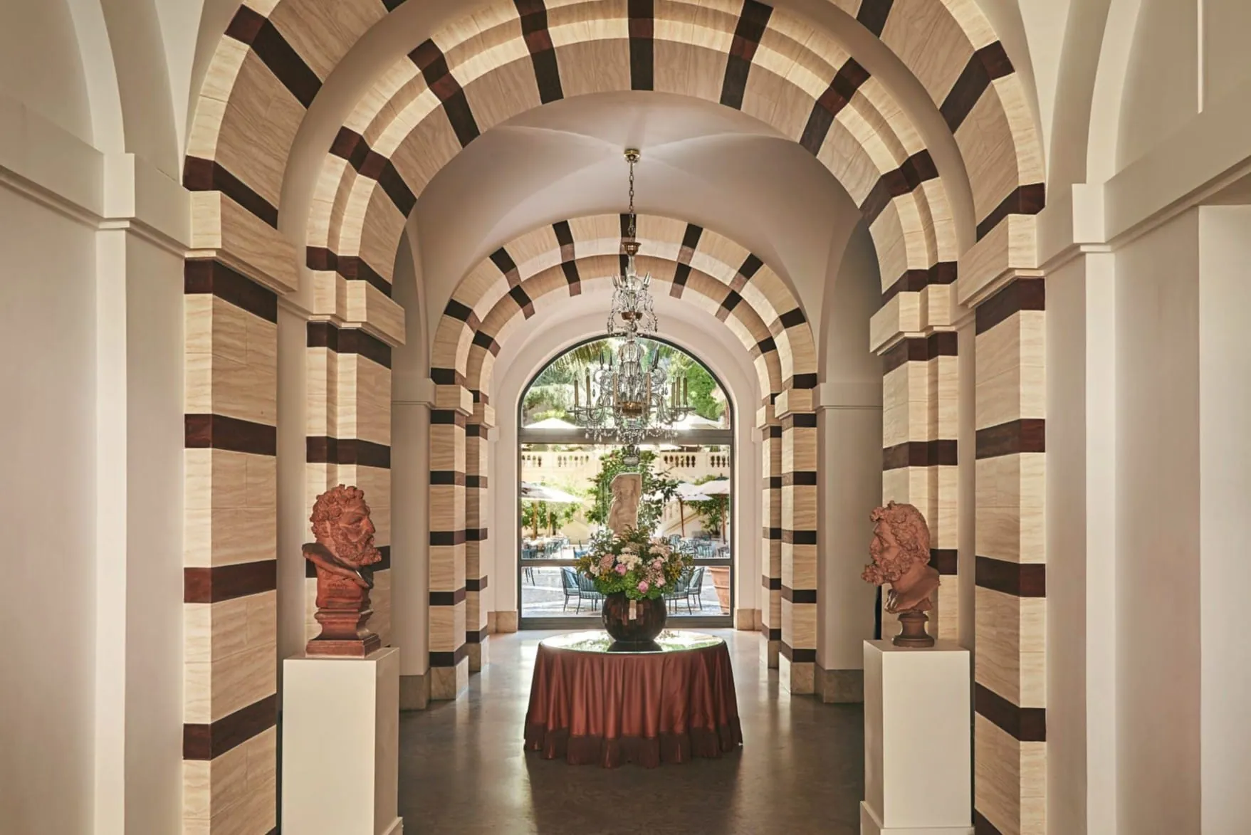 Klassische römische Rundbögen mit kontrastreichen Streifen rahmen den Blick in den „Secret Garden“ des Hotels de Russie. Antike Terrakotta-Büsten auf weißen Säulen und ein Kristallleuchter unterstreichen das Flair des Entrees.