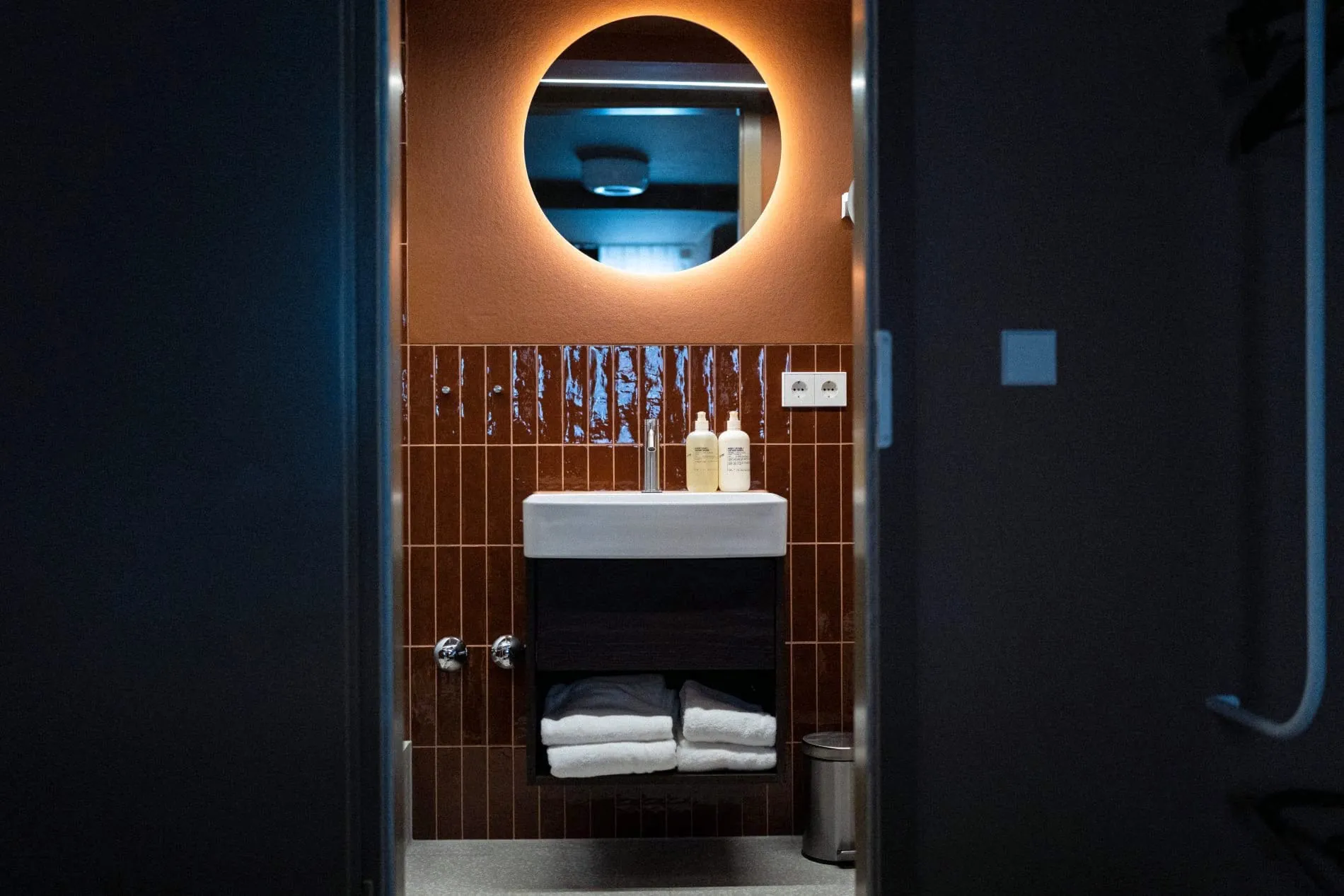 Die Badezimmer im Maison Meissner spiegeln das Farbkonzept des Hauses wider. Dunkelrote Fliesen harmonieren mit dem warm hinterleuchteten Rundspiegel über dem Waschtisch.