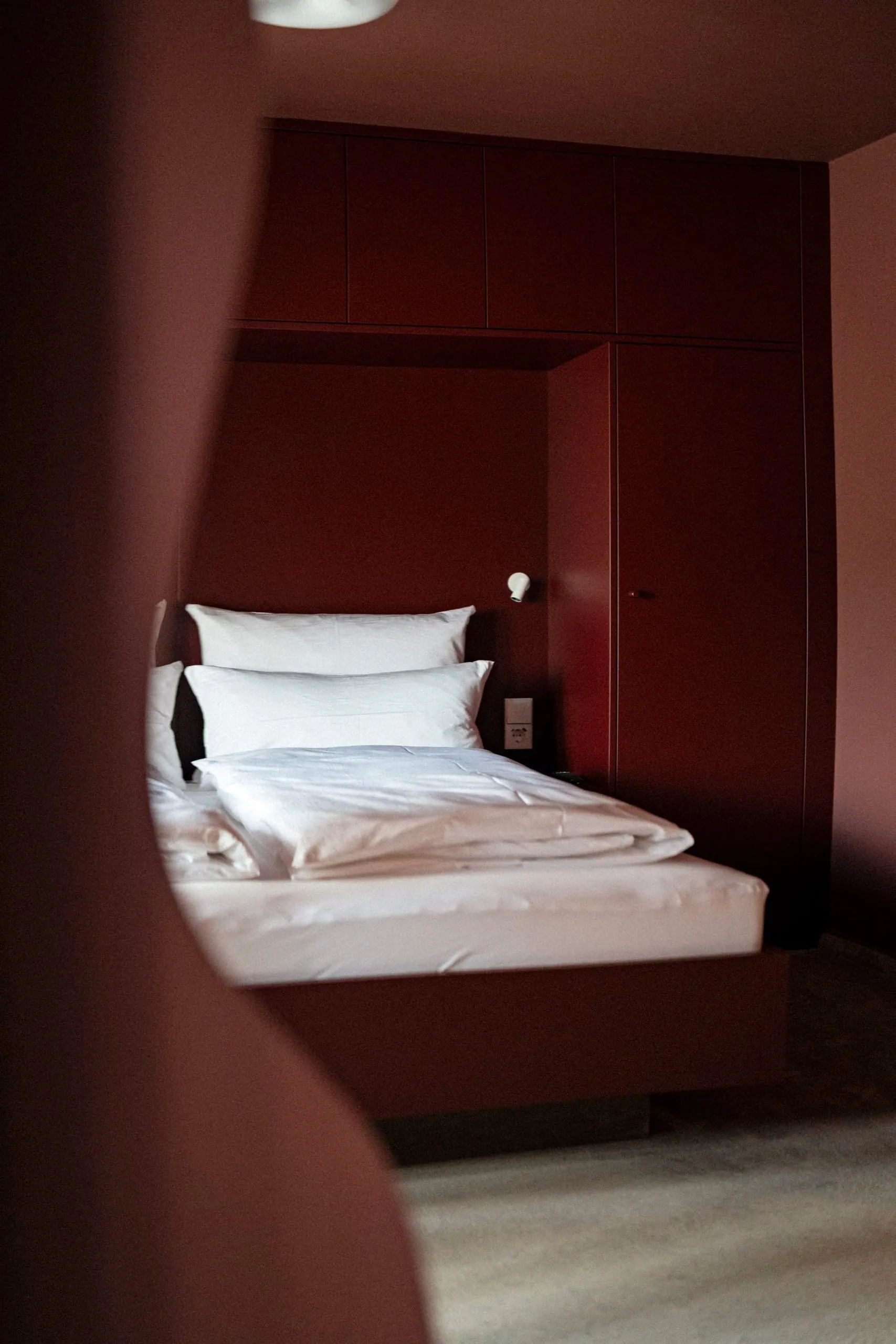 In den kleineren Zimmern des Hotels nutzen maßgefertigte Einbauten eines Allgäuer Schreiners den Raum optimal aus. Das Bett schmiegt sich nahtlos in die dunkelrote Schrankwand ein.