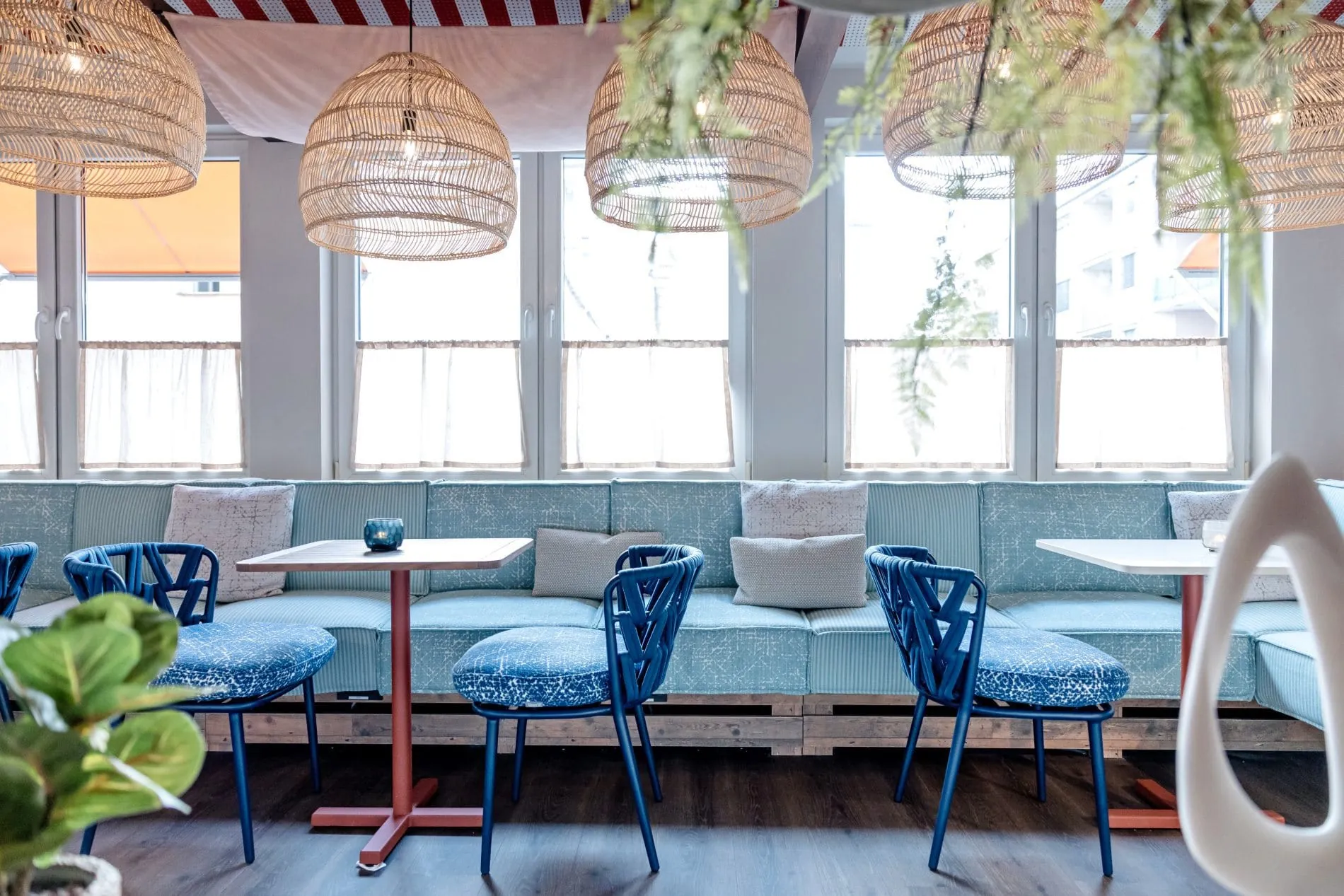 Der angeschlossene Wintergarten erweitert die Lounge um einen hellen Sitzbereich. Hellblaue Polster, Seilstühle, Rattanleuchten und ein Markisentuch erinnern an Streetfood-Märkte und urbane Außenräume.
